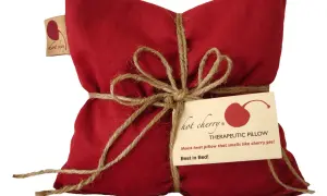 Cuscino rosso con noccioli di ciliegia, legato con spago. Un'etichetta recita "Hot Cherry Therapeutic Pillow". Benefici rilassanti.