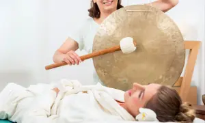 Donna sdraiata su lettino da massaggio, avvolta in un accappatoio, mentre un'altra donna suona un gong. Musica rilassante per massaggi.