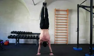 Uomo esegue flessioni per spalle in un centro fitness, con bilancieri e spalliera svedese sullo sfondo.