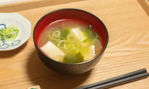 Una ciotola di brodo di miso fumante con tofu e cipollotti, accompagnata da una piccola ciotola di cipollotti affettati e bacchette.