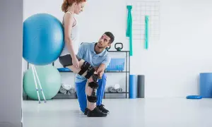 Chinesiologo cosa cura: riabilitazione ginocchio con tutore e palla. Terapista assiste paziente in esercizi.