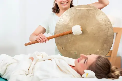 Donna sdraiata su lettino da massaggio, avvolta in un accappatoio, mentre un'altra donna suona un gong. Musica rilassante per massaggi.