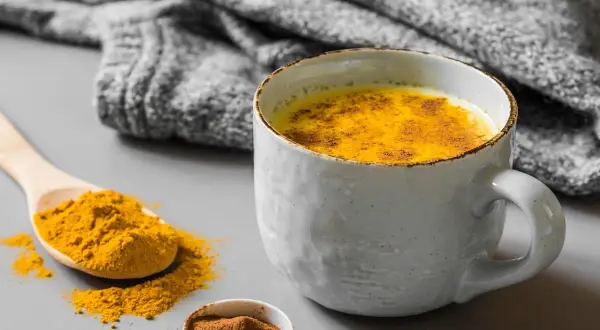 Un latte dorato speziato, perfetto cosa bere al posto del caffè. Curcuma e cannella in una tazza calda, con una coperta grigia sullo sfondo.