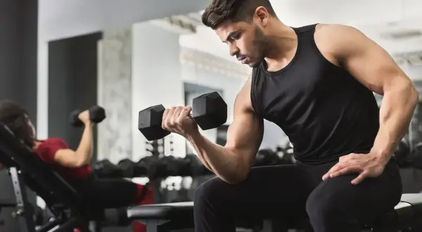 Uomo concentrato esegue esercizi con manubri in palestra. A sinistra, una donna fa lo stesso su una panca inclinata.