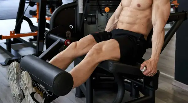 Uomo scolpito esegue un **circuito gambe e glutei** in palestra, concentrato sull'allenamento dei quadricipiti con la macchina.