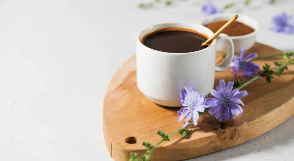 Tazza di caffè di cicoria scuro su tagliere di legno, decorata con fiori di cicoria viola.