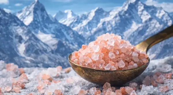 Cristalli di sale rosa su un cucchiaio, con montagne innevate sullo sfondo. Questi cristalli di sale a cosa servono? Per insaporire i cibi e per il benessere.