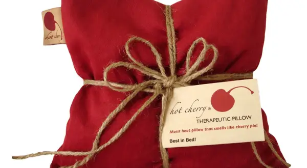 Cuscino rosso con noccioli di ciliegia, legato con spago. Un'etichetta recita "Hot Cherry Therapeutic Pillow". Benefici rilassanti.