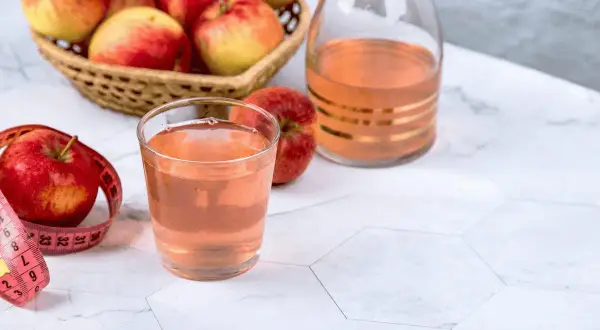 Un bicchiere di aceto di mele, mele fresche in un cesto e una bottiglia decorata. Un metro da sarta rosa avvolge una mela, suggerendo un percorso di benessere.
