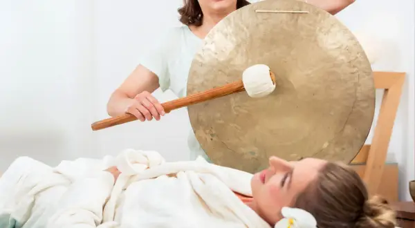 Donna sdraiata su lettino da massaggio, avvolta in un accappatoio, mentre un'altra donna suona un gong. Musica rilassante per massaggi.