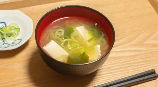 Una ciotola di brodo di miso fumante con tofu e cipollotti, accompagnata da una piccola ciotola di cipollotti affettati e bacchette.