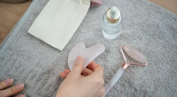 Mano che tiene una pietra gua sha rosa, accanto a un rullo di quarzo e un siero. Utile per il benessere, ma attenzione alle gua sha controindicazioni.