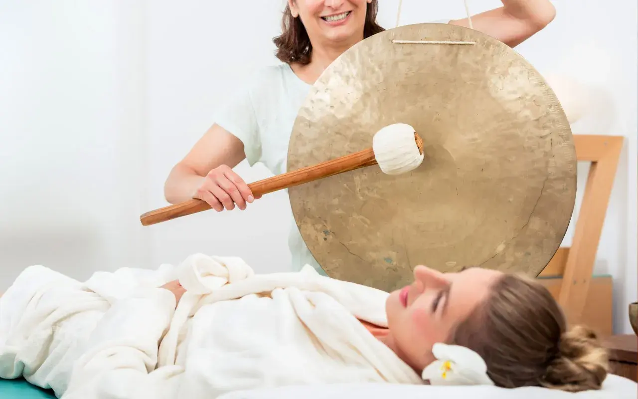 Donna sdraiata su lettino da massaggio, avvolta in un accappatoio, mentre un'altra donna suona un gong. Musica rilassante per massaggi.