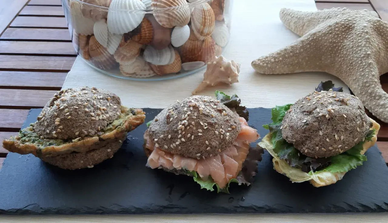 Tre panini low carb ricetta: uno con pesto, uno con salmone affumicato e insalata, uno con formaggio e insalata. Decorazioni marine sullo sfondo.