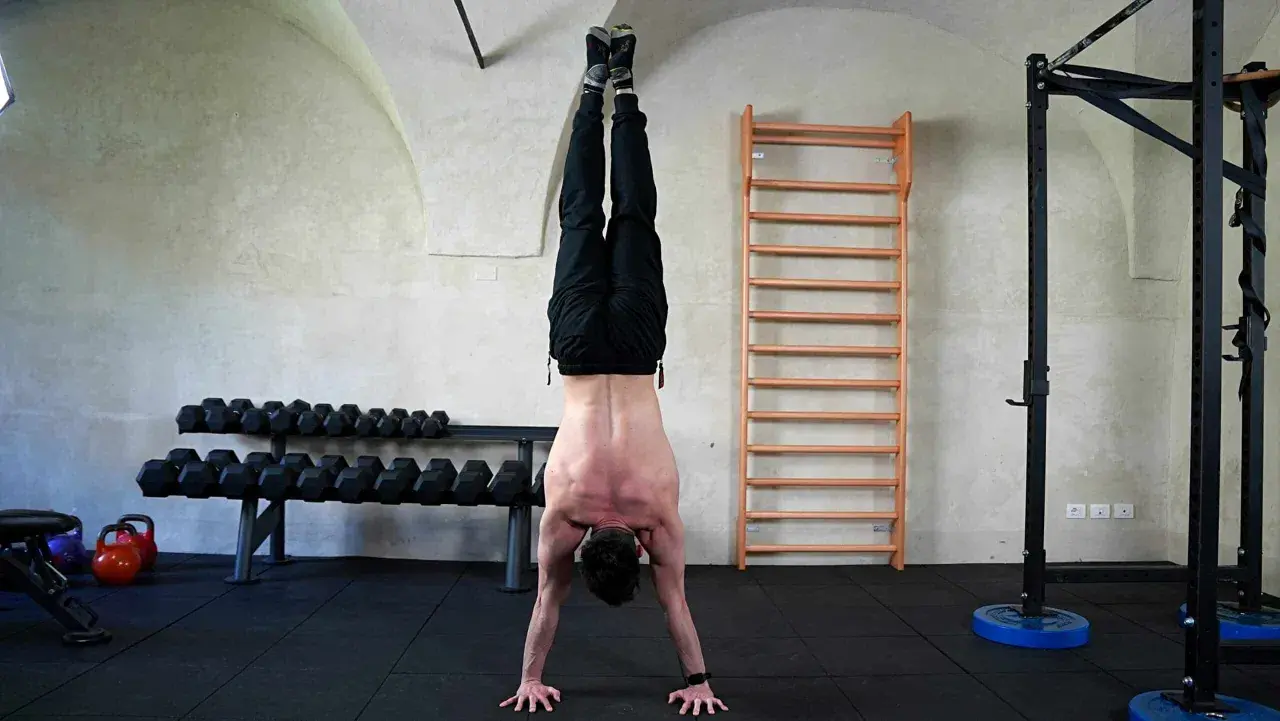 Uomo esegue flessioni per spalle in un centro fitness, con bilancieri e spalliera svedese sullo sfondo.