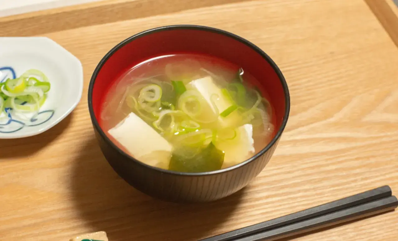 Una ciotola di brodo di miso fumante con tofu e cipollotti, accompagnata da una piccola ciotola di cipollotti affettati e bacchette.