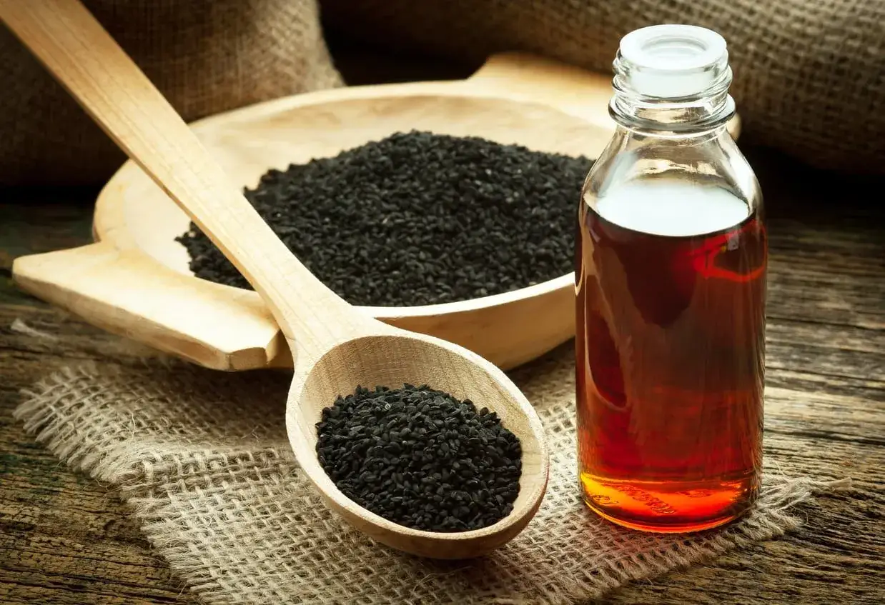 Semi di cumino nero in un cucchiaio di legno e una bottiglia d'olio ambrato. Scopri come mangiarli per il benessere.