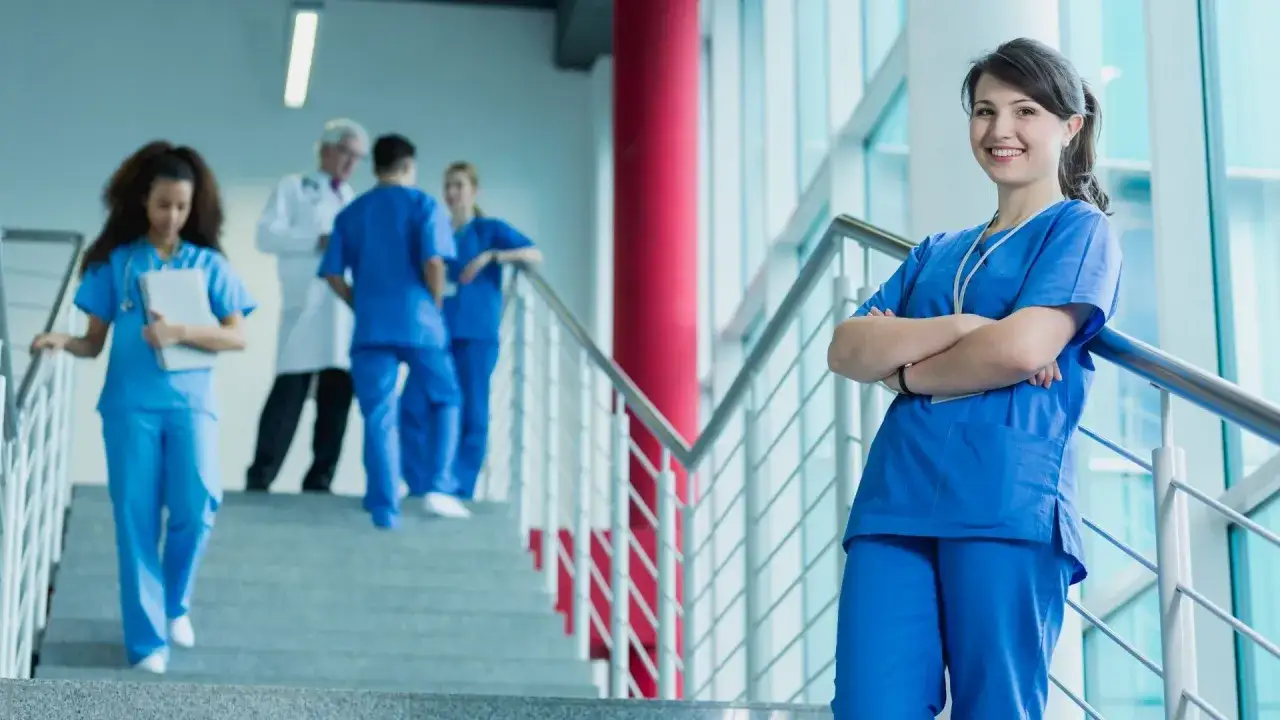 Giovane donna sorride con le braccia conserte, un'ispirazione per chi vuole diventare fisioterapista. In sottofondo, studenti e medici in corridoio.
