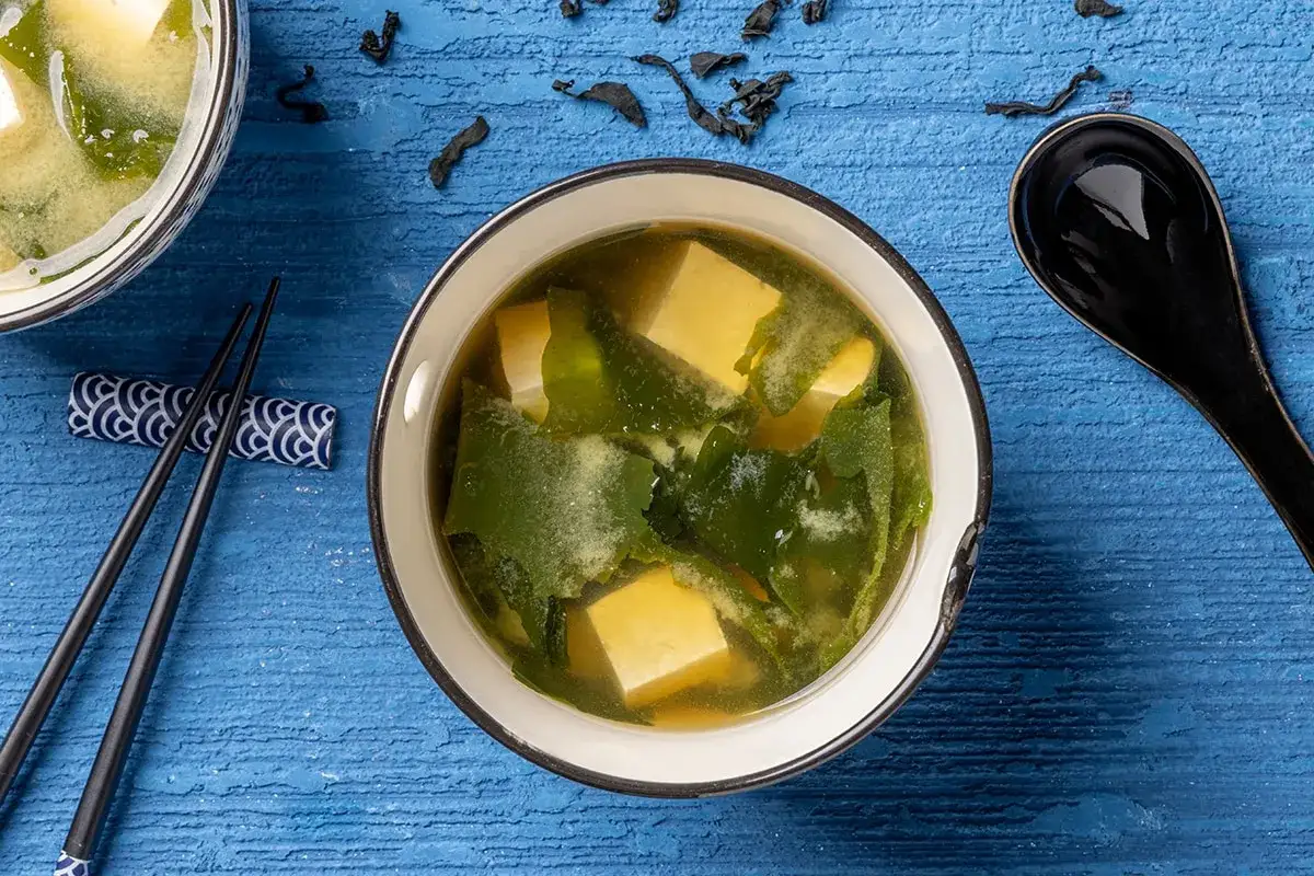 Un fumante brodo di miso con cubetti di tofu e alghe verdi, pronto per essere gustato con bacchette e cucchiaio nero.