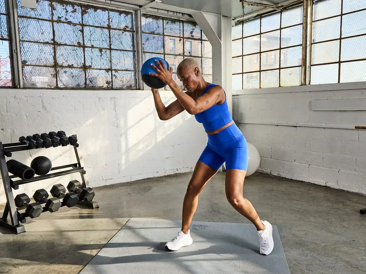 Donna esegue esercizi con la palla per dimagrire in palestra, con manubri e foam roller sullo sfondo.