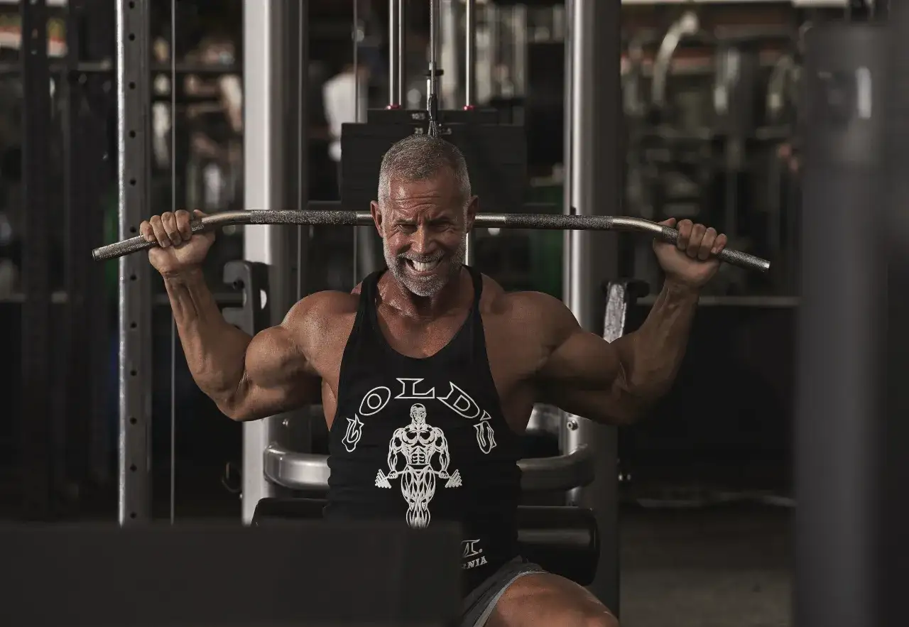 Uomo muscoloso con barba grigia esegue esercizi con bilanciere in palestra, sudato e concentrato.
