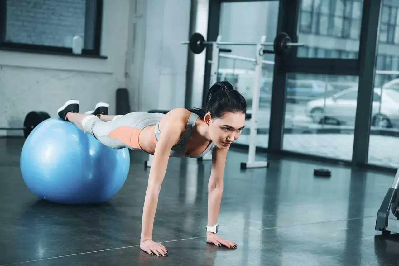 Donna esegue addominali con la palla, con i piedi appoggiati sulla palla da fitness blu, in una palestra luminosa.
