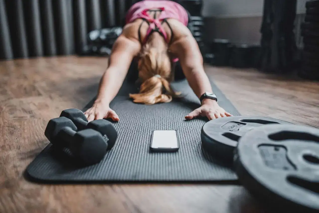 Donna in posa yoga sul tappetino, con manubri e pesi. Il telefono segna i 30 secondi di recupero tra le serie.