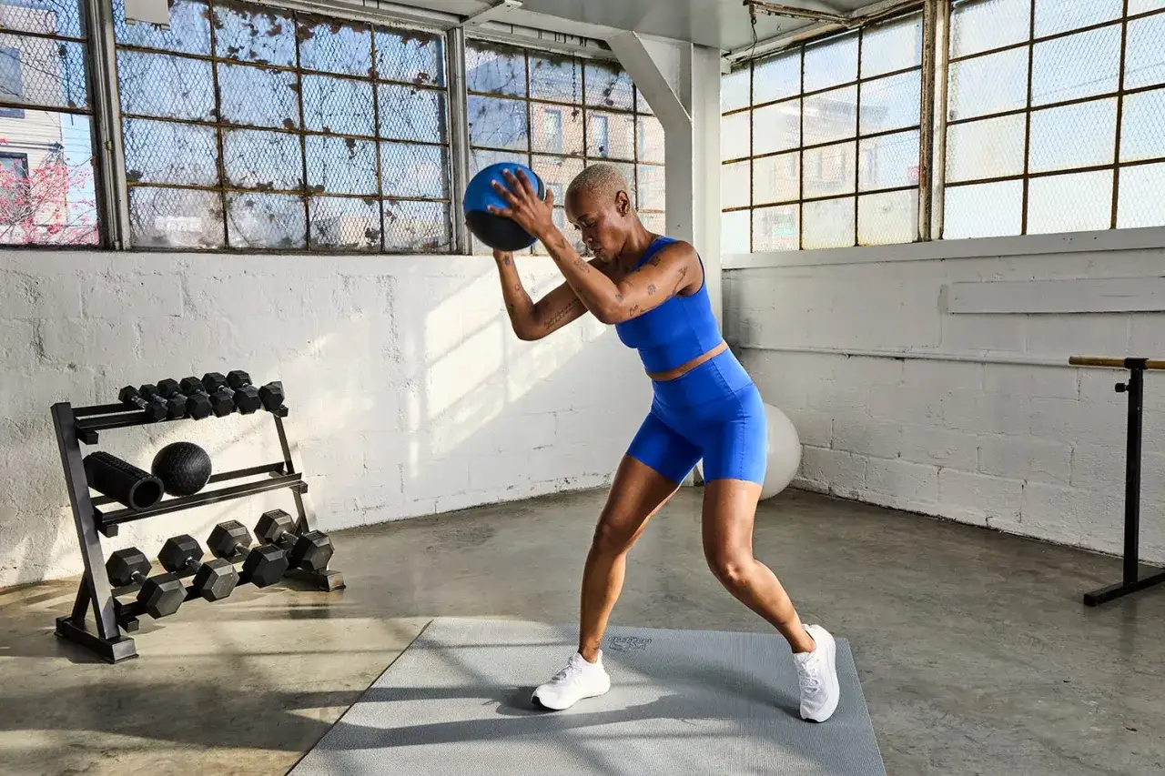 Donna esegue esercizi con palla medica in palestra. Allenamento intenso con pesi e attrezzi sullo sfondo.