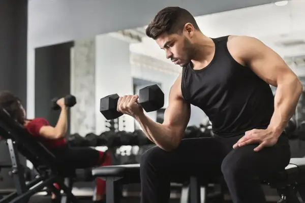 Uomo concentrato esegue esercizi con manubri in palestra. A sinistra, una donna fa lo stesso su una panca inclinata.