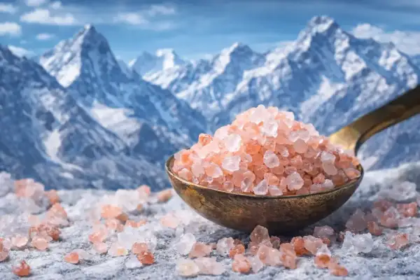 Cristalli di sale rosa su un cucchiaio, con montagne innevate sullo sfondo. Questi cristalli di sale a cosa servono? Per insaporire i cibi e per il benessere.